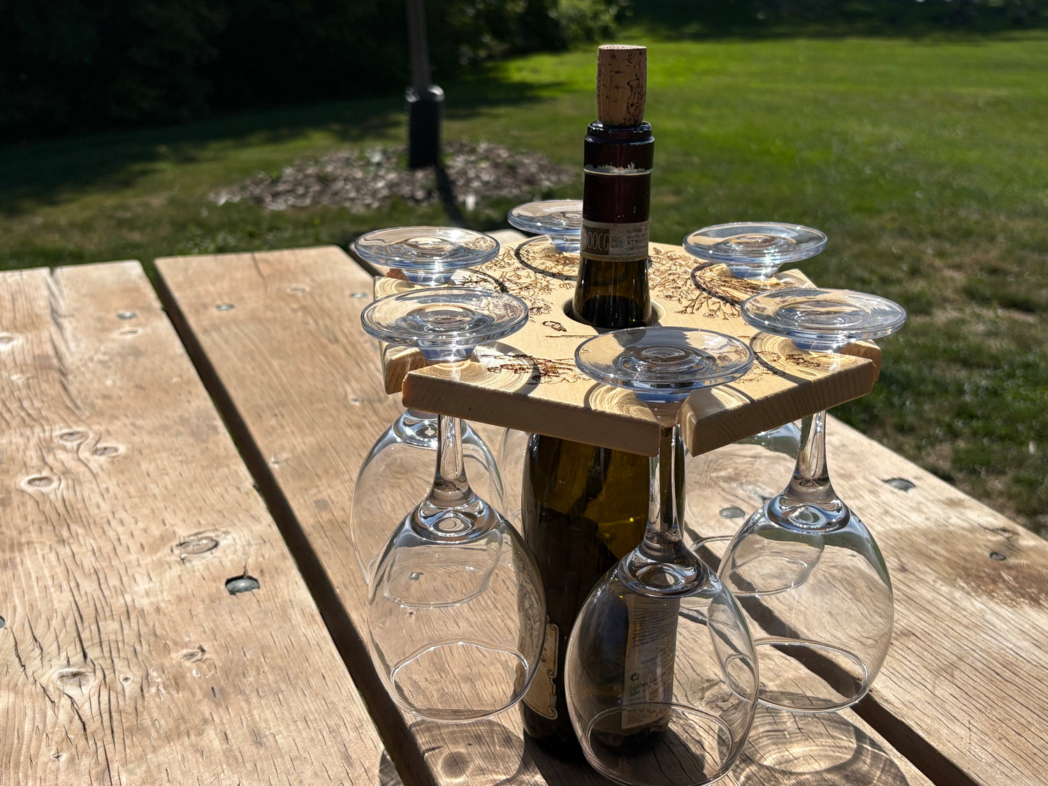 Wine bottle with corkscrew and glasses on a wooden table outdoors with our 6-glass wine caddy atop and six glasses hanging off it.